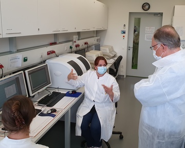 Eine Laborantin sitzt vor Bildschirmen und Tastaturen in einem Laborraum und erklärt dem vor ihr stehenden Ministerpräsidenten wie eine PCR-Untersuchung abläuft. Beide tragen weiße Schutzkittel und eine Mund-Nasen-Bedeckung.
