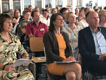 Ein Forum von vielen Menschen lauscht einem Vortrag. Sie sitzen in Reihen mit dem Blick leicht nach rechts gerichtet. Im Vordergrund von links nach rechts: Barbar Woltmann, Miriam Staudt, Eberhard Haunhorst