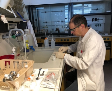 Ein LAVES-Mitarbeiter sitzt an einem Labortisch vor einem Waschbecken. Er trägt einen weißen Kittel und eine Schutzbrille sowie Einmal-Handschuhe. Neben ihm auf dem Tisch diverse Laborutensilien.
