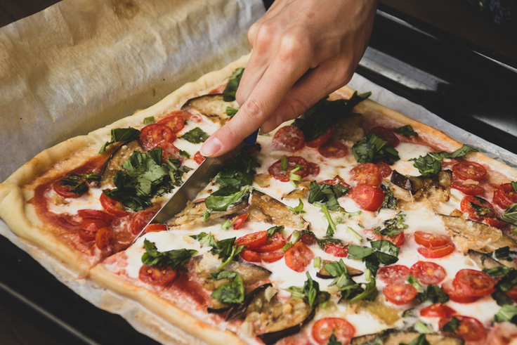 Ein Messer schneidet einen aufgebackenen Pizzateig auf dem Rucola, Tomaten und Mozzarella liegt.