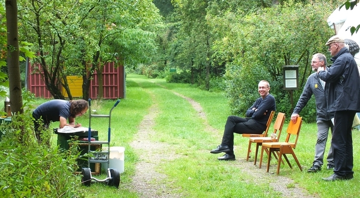 Ein Prüfling lehnt über einer grünen Kiste mit einem Bienenvolk. Ihm gegenüber beobachten drei Prüfer das Geschehen.