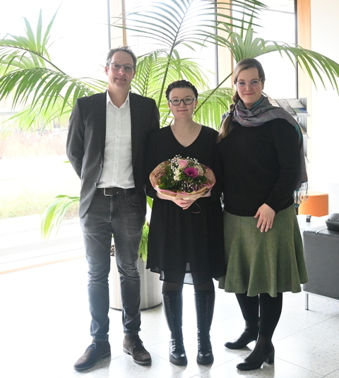 Die drei Menschen stehen vor einem großen Farn. Vivien Kassandra Weihreich hält einen bunten Blumenstrauß in der Hand.