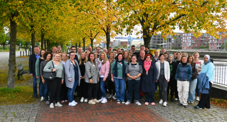 Gruppenbild der Willkommensveranstaltung am Hafen in Oldenburg unter Bäumen mit gelben und grünen Blättern.