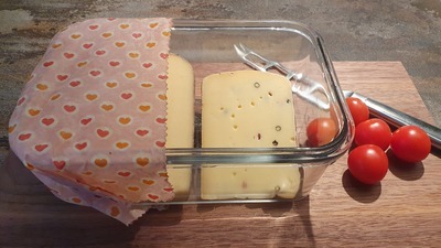 Ein rechteckige Glasschale mit zwei Stücken Käse ist zur Hälfte mit einem Bienenwachstuch abgedeckt. Daneben liegen ein paar Tomaten und ein Käsemesser.