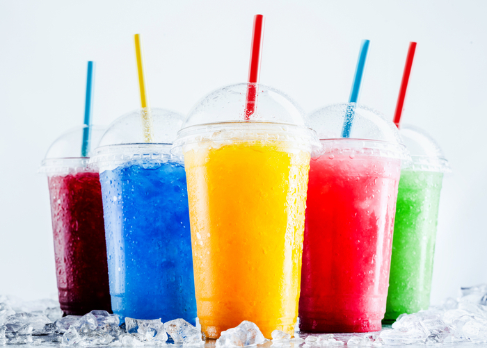 Gefrorene Frucht-Slush-Getränke in Plastikbechern zum Mitnehmen mit Deckel und Trinkhalmen, die auf einer kalten Metalloberfläche mit verstreuten Eiswürfeln kühlen