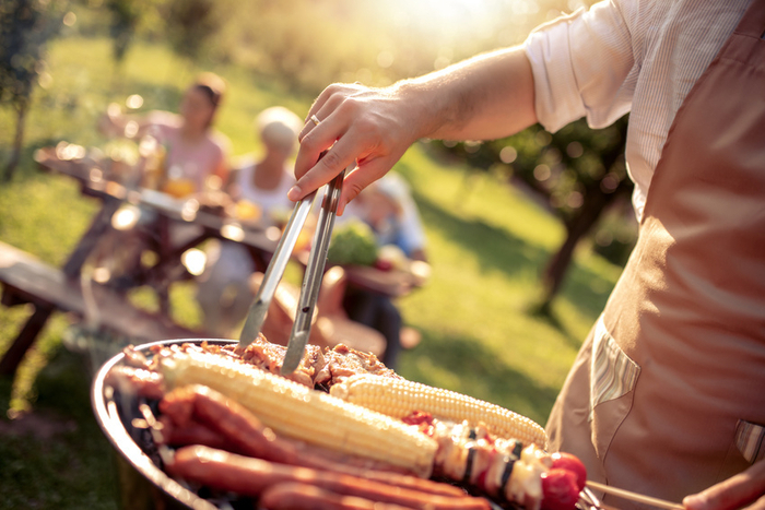 Eine Person steht am Grill im Garten, im Hintergrund eine Familie sitzend am Tisch erkennbar